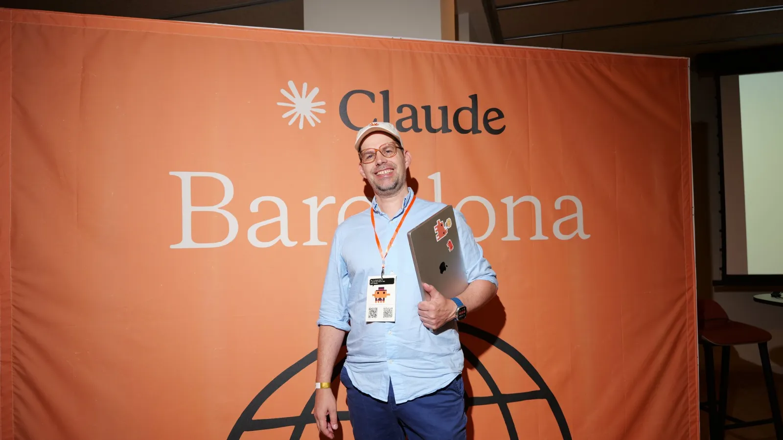 Florian Steiner mit Laptop vor der orangefarbenen Wand mit der Aufschrift Claude Barcelona im Claude Community House